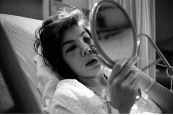 Black and white photograph of a woman in a hospital bed looking at herself in a handheld mirror.